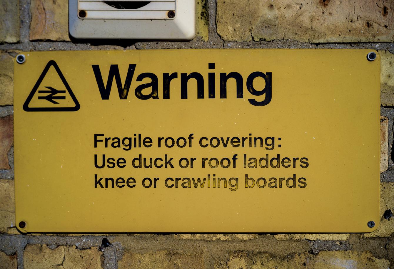 A black-on-yellow sign reading &ldquo;Warning - Fragile roof covering: Use duck or roof ladders knee or crawling boards&rdquo;.