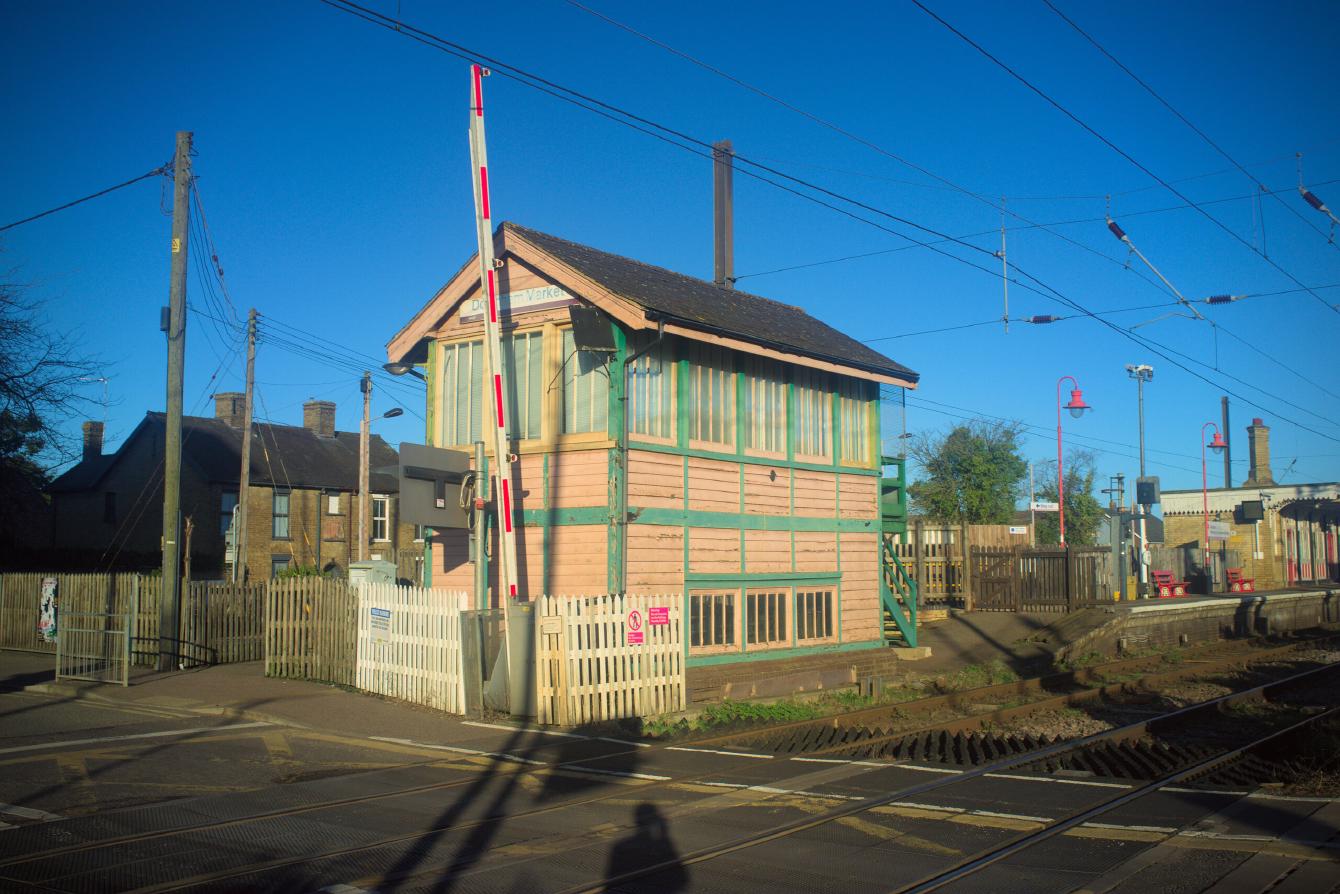 The signal box at Downham Market. It has a green wooden frame filled with cream-coloured slats. In the foreground is a level crossing barrier