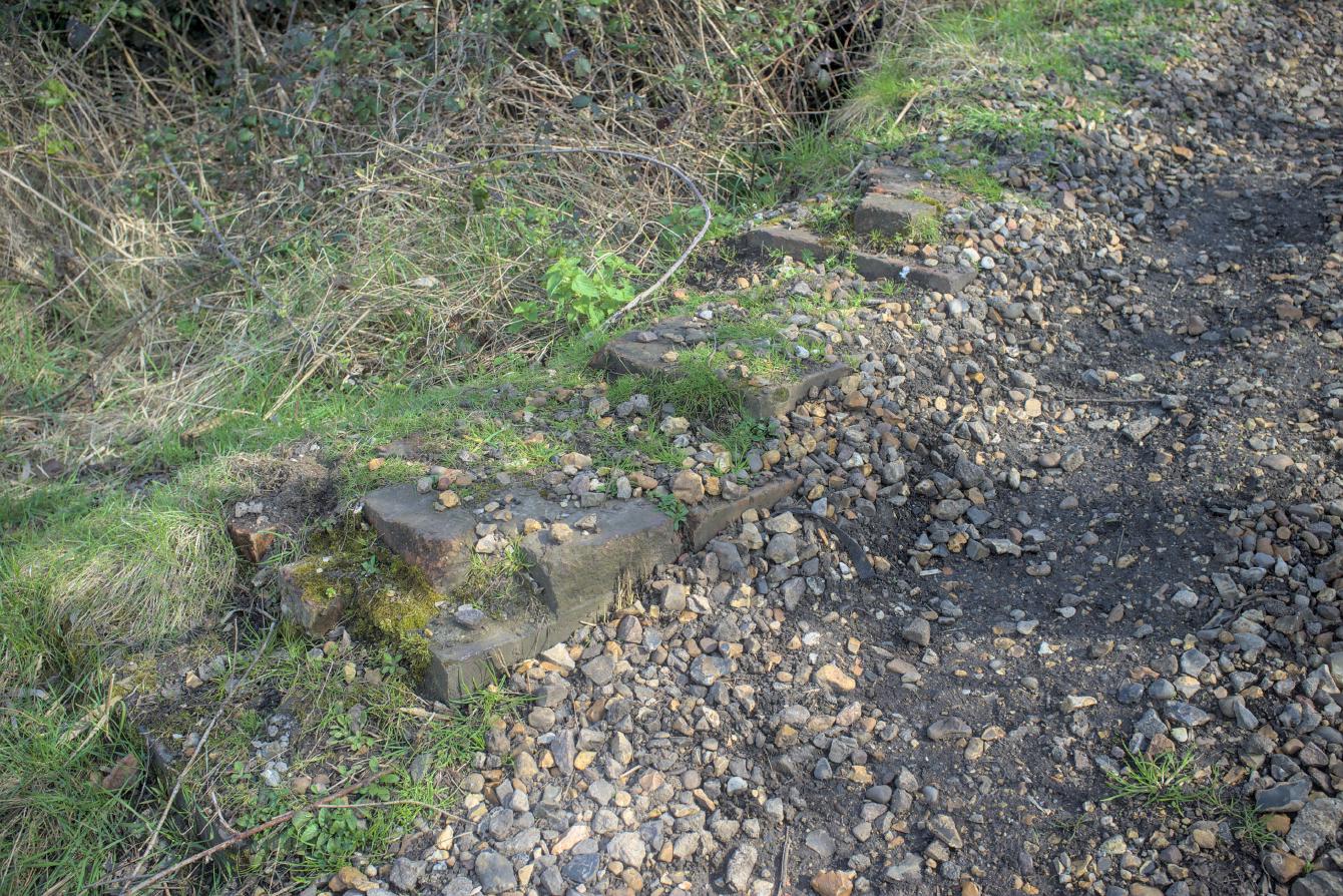 A closeup of a collapsed wall; the top has disappeared, with remaining brickwork embedded in the ground showing the path it would have taken.