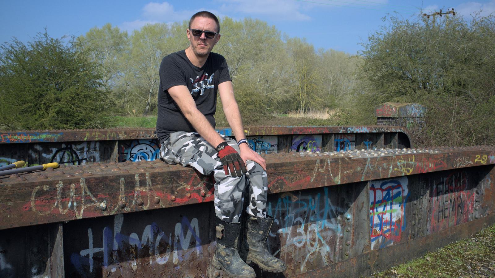 An older me sitting on the same bridge as in the first photo, in combat trousers and a tshirt and rigger boots; not much has changed.