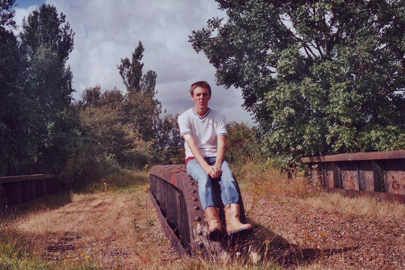 Former me, dude in his 20s with rigger boots and jeans and a white t-shirt, sitting on a rusty iron beam of an old railway bridge.