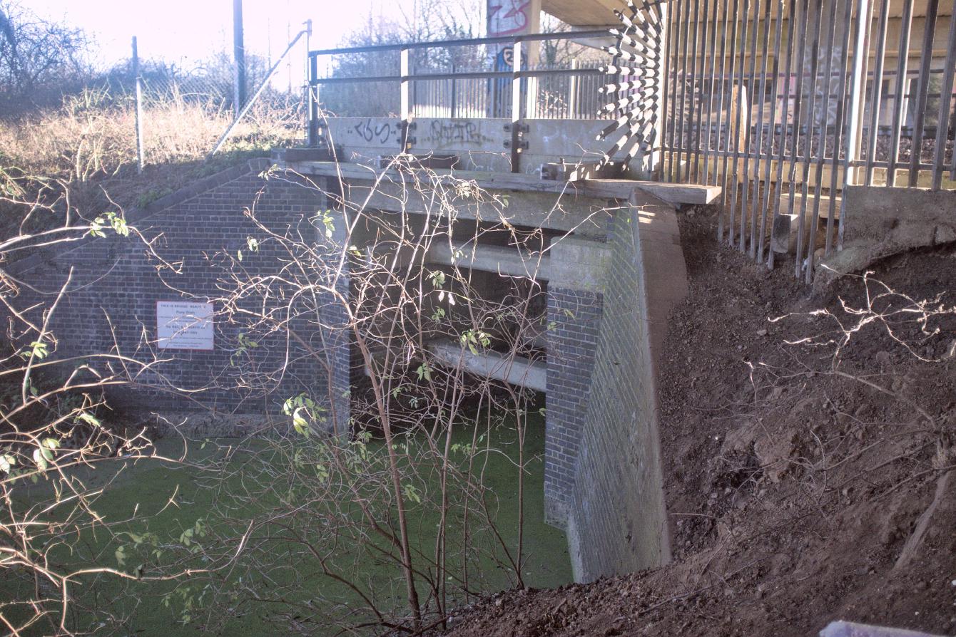 A tiny railway bridge over a green-covered drain