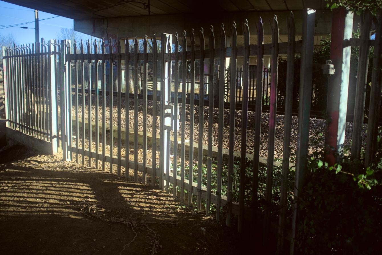 A steel security gate; it is mostly made of vertical galvanised steel slats, and has spikes on the top to prevent climbing. A single (active) railway track is visible behind it.