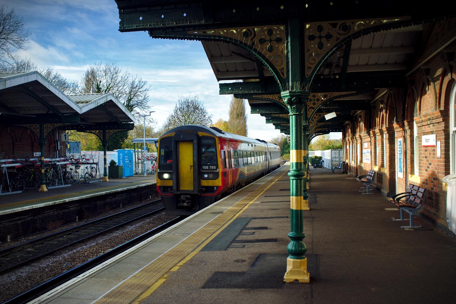 A class 158 arriving at platform 2 at March station. It is a two-car diesel multiple unit train. A low sun illluminates the scene from the left, and in between the canopies of the platforms is a blue sky.