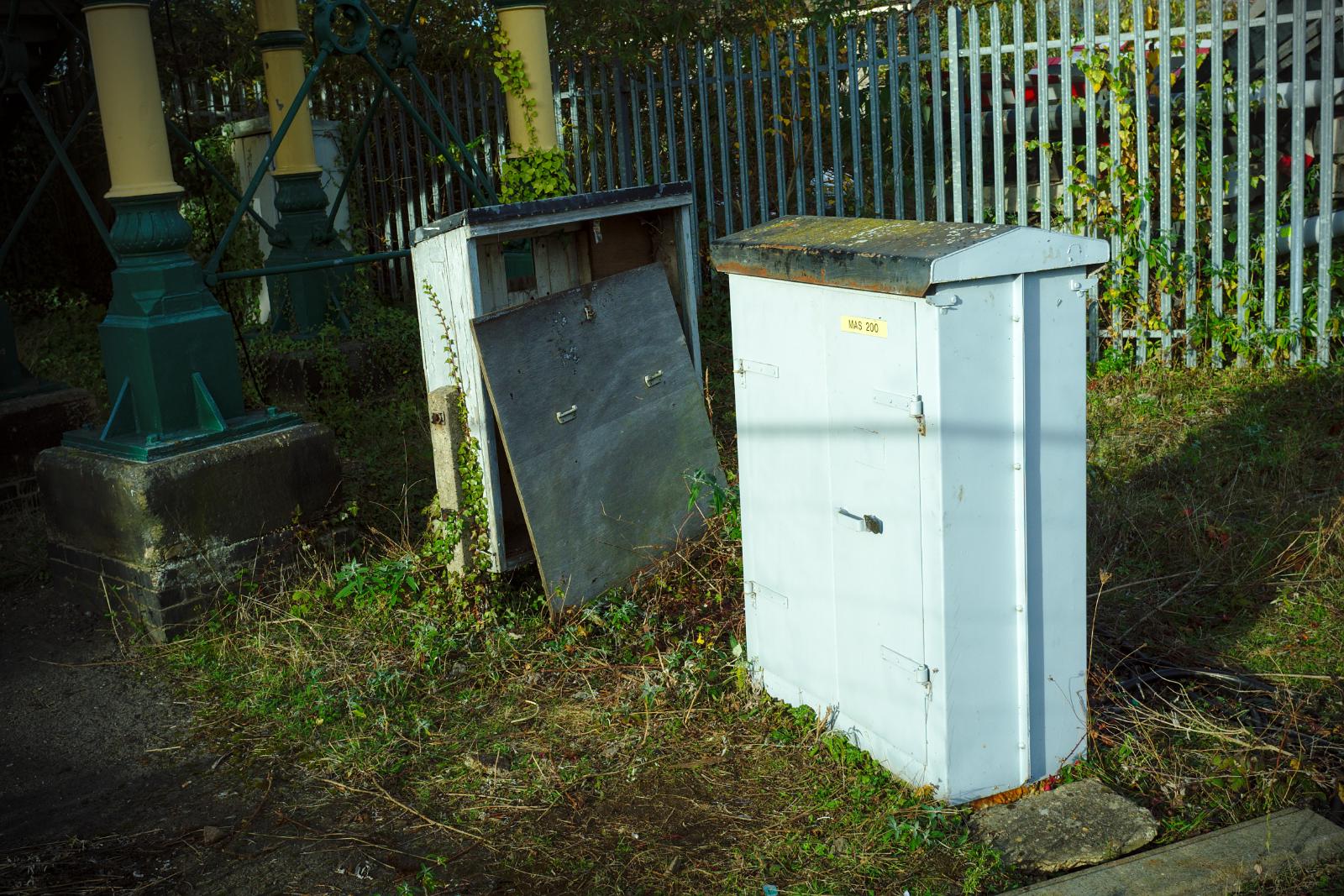 Some old electrical/signalling equipment boxes. One has a door present, but merely leaning against the box. In the background are the pillars of the footbridge.