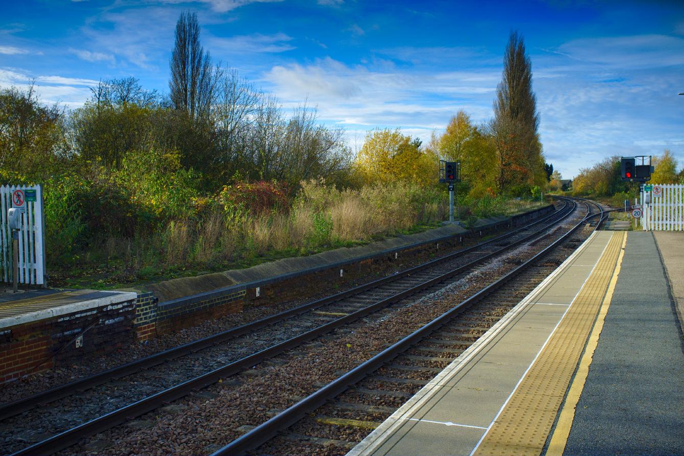 A view of the very end of March railway station platform 1, viewed across the tracks from platform 2. A white fence limits how far customers may walk, but the platform continues for some distance. The unused part is overgrown. A signal, in use and indicating red, sits in the disused part.