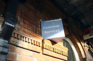A triangular Customer Services sign attached to some brickwork. It is blue and a little faded. Anti-climbing spikes protrude from the top.