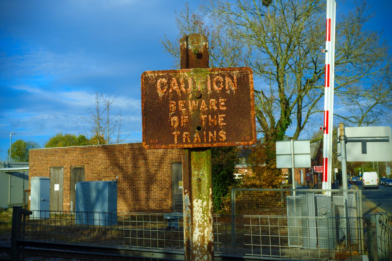 An old sign saying &ldquo;Caution: Beware of the trains&rdquo; in capital letters. The sign is rusted, but the text is clearly visible. You can just make out some text saying &ldquo;LNER&rdquo; and &ldquo;0.161&rdquo; on the bottom corners. A level crossing barrier is visible in the background.