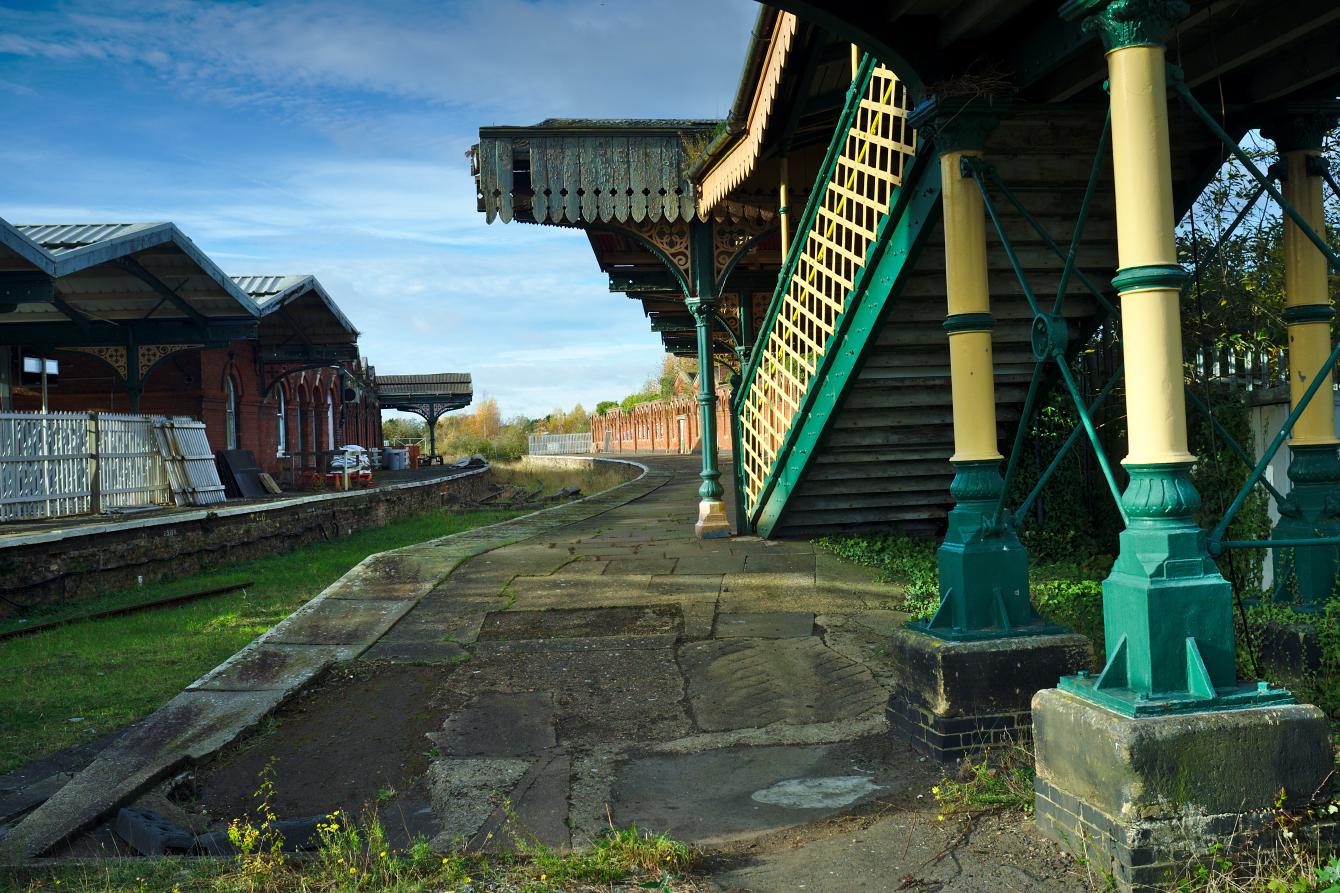 A view down two disused platforms at March station. On the right are the pillars of the section of the footbridge that used to lead down here. Part of the original canopies are visible on both platforms. The area between the platforms where the track was is grassy, but well-kept.