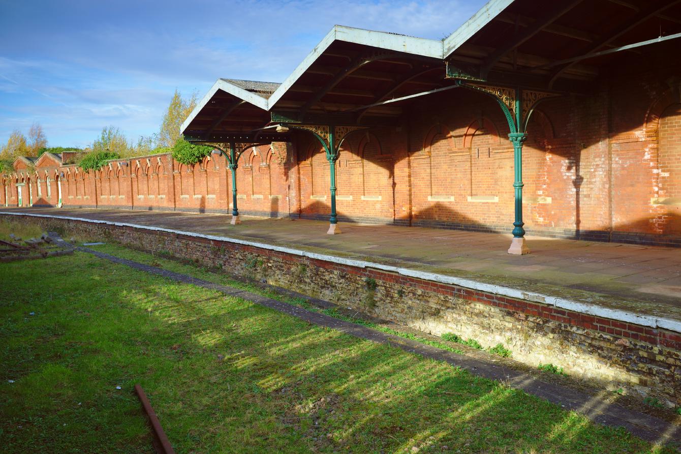 A view of one of the platforms from the picture above. The section of canopy is visible; it has the same triangular pattern as the canopies on the in-use section of the station. Behind the platform are brick walls in a very nice ornamented Victorian style. Well-kept grass on the trackbed is visible in the foreground.