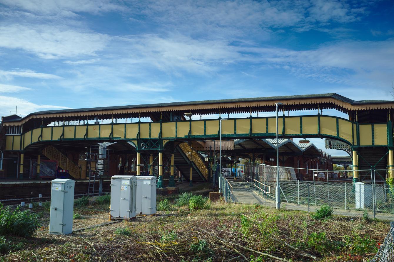 A view of March station footbridge. It is constructed in iron, and is cream-coloured with green accents. It is large enough to span two sets of tracks, though there is a footpath under the span on the right instead of railway tracks (which have been removed). It is a sunny day with blue skies.