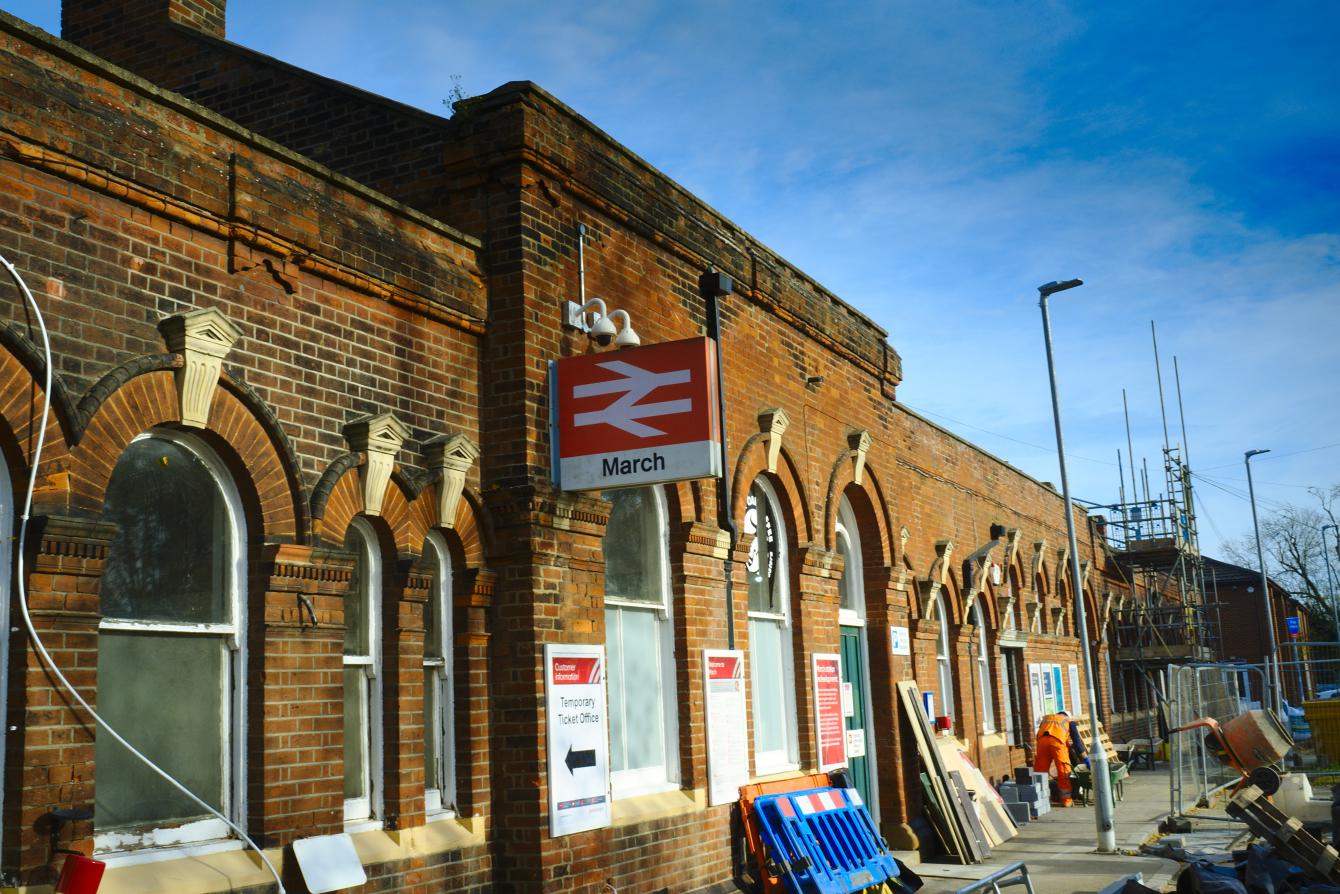 The outside of March railway station building. Various construction clutter sits around. The building is brick and ornamented ina very Victorian style. There is a sign with a British Rail double-arrow with &ldquo;March&rdquo; in black-on-white underneath.