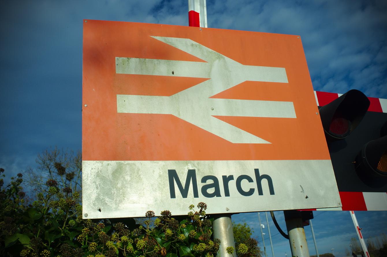 The station sign next to the level crossing at March station. A double arrow symbol, white on flame red, sits above black-on-white text saying &ldquo;March&rdquo;.