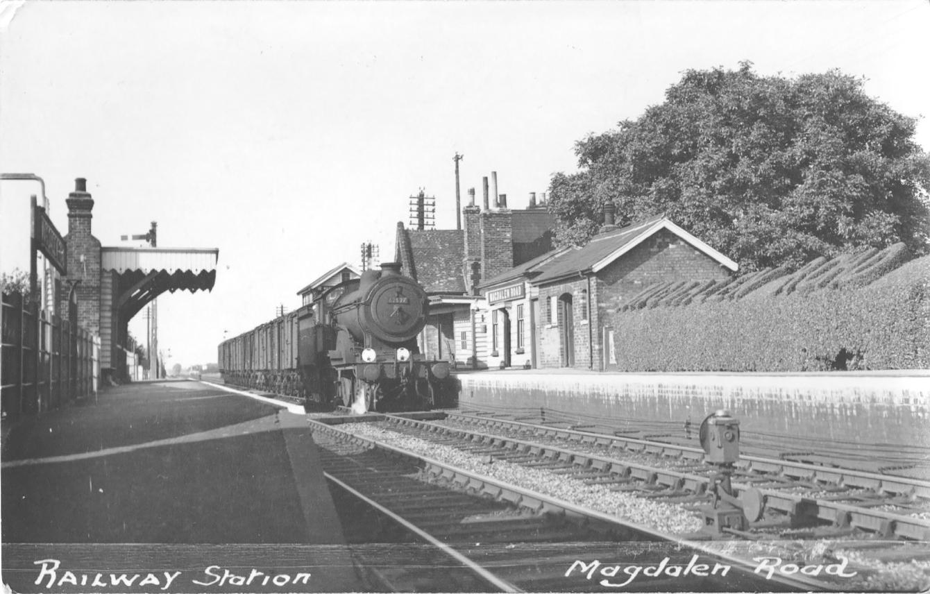 A steam locomotive passes through Magdalen Road. In the right of the photo, a hedge has the text &ldquo;Magdalen Road&rdquo; cut into its top.