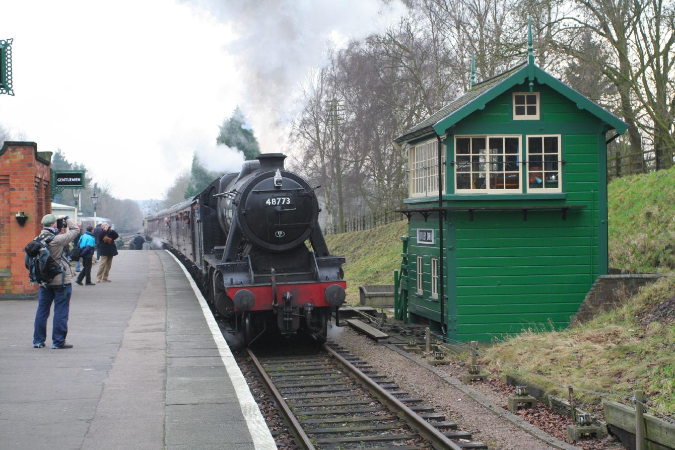 The signal box at Rothley; it is a wooden signal box painted green, with cream details. A steam locomotive sits next to it.